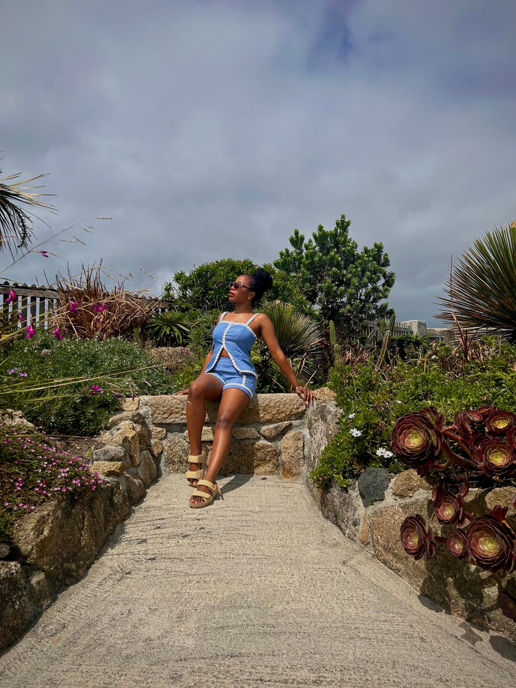 Jasmine Lubi seated in a coastal Cornish garden, sunglasses on, gaze turned up and away