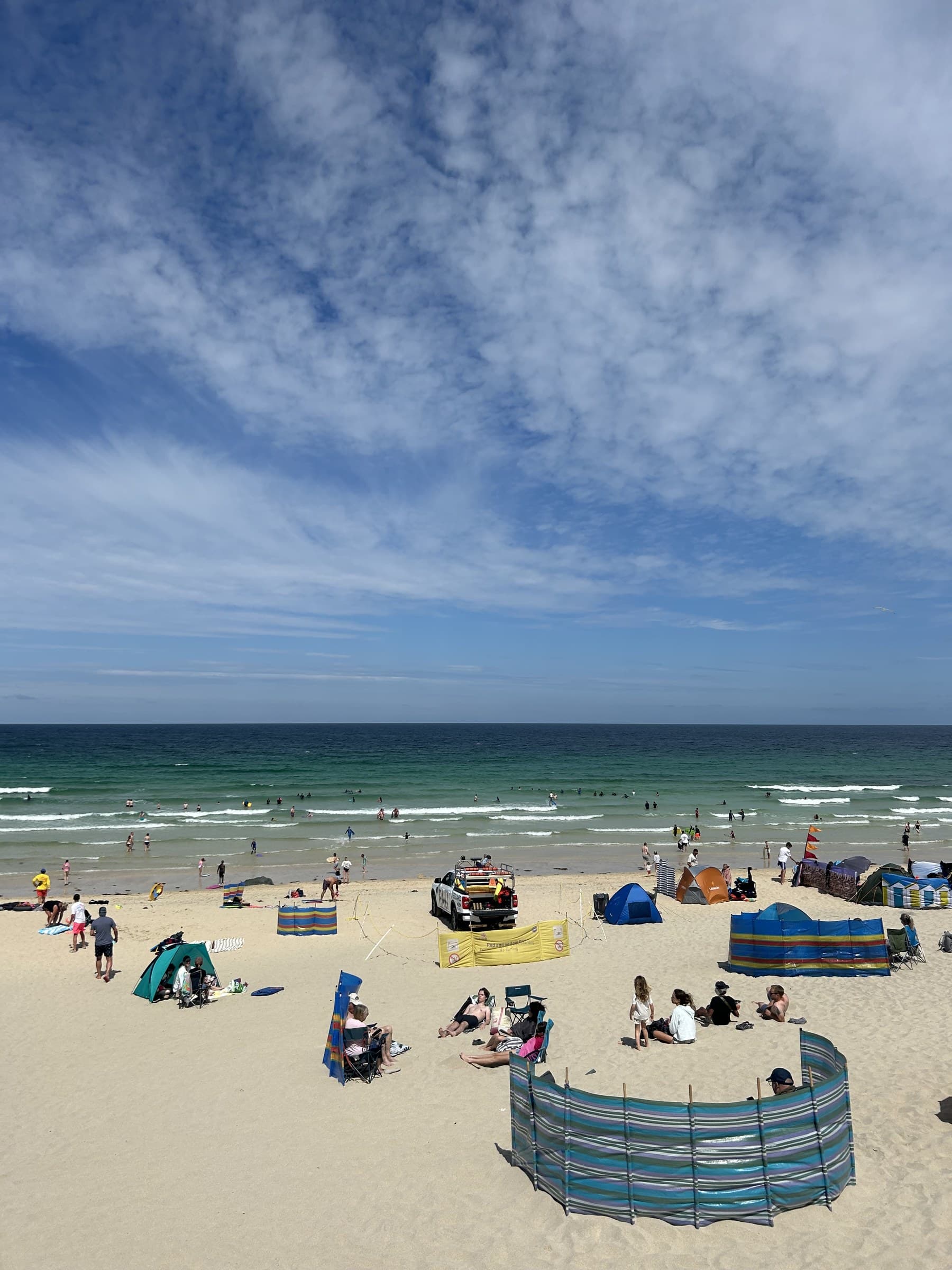 A busy, clean Cornish beach under a wide sky, windbreaks and families dotted across the sand