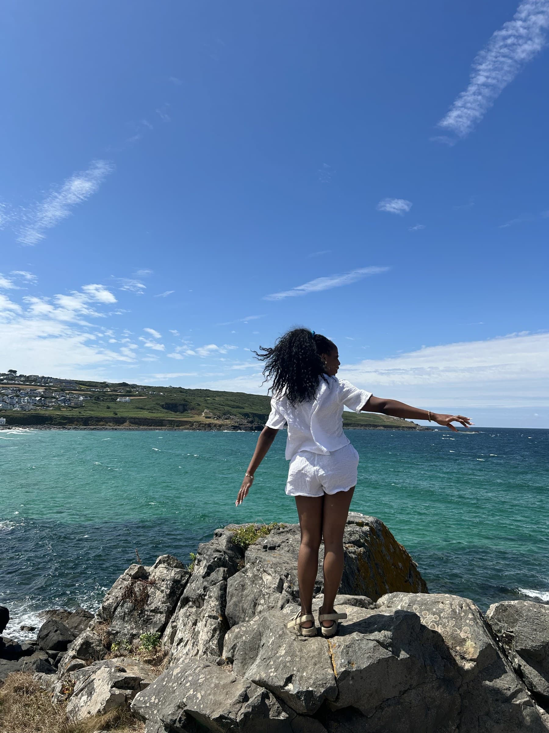 Jasmine Lubi standing on a coastal rock at The Lizard, arms out, looking out to sea