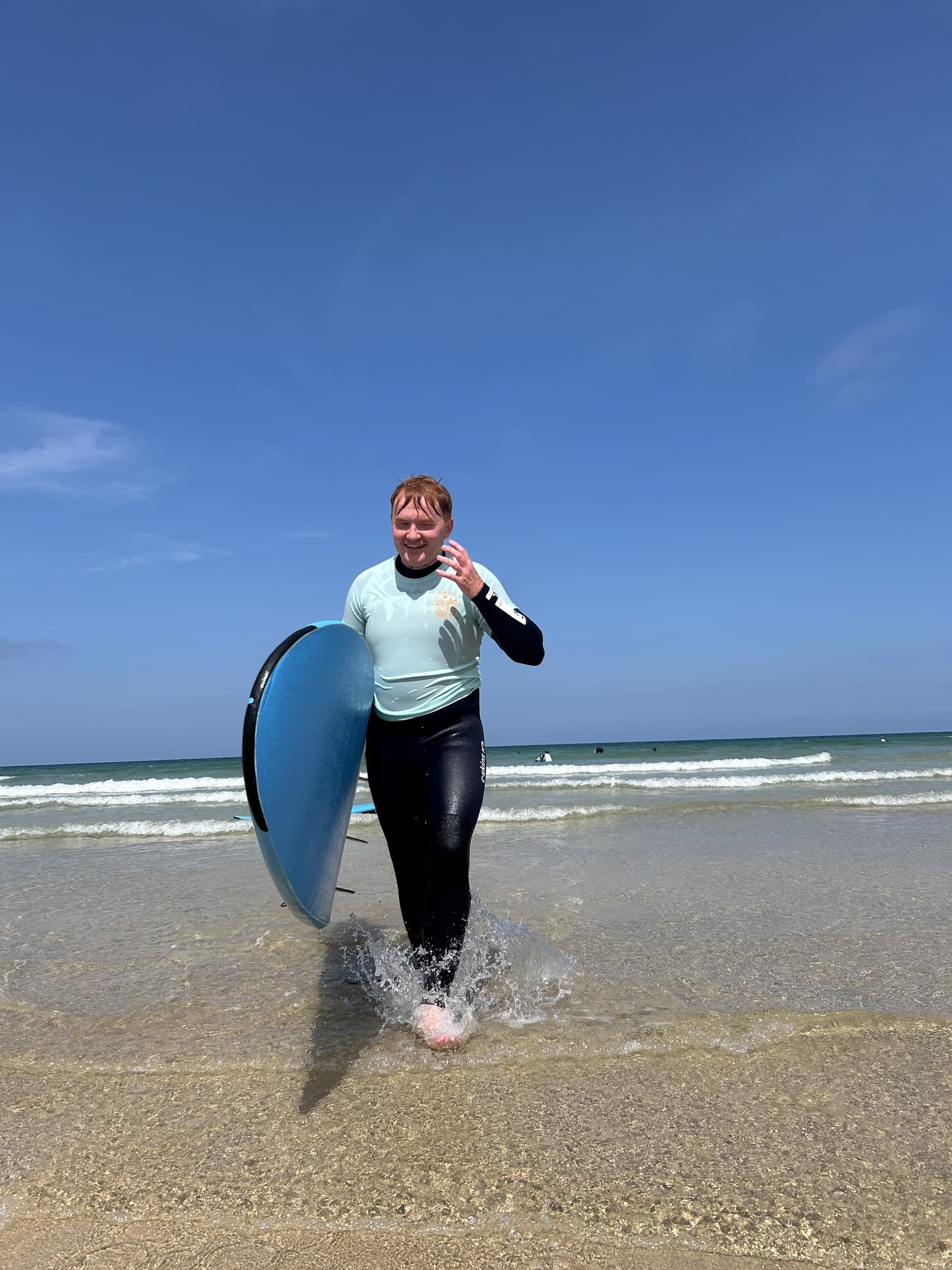 Jasmine's partner walking out of the sea after his first surfing lesson, board under his arm, laughing